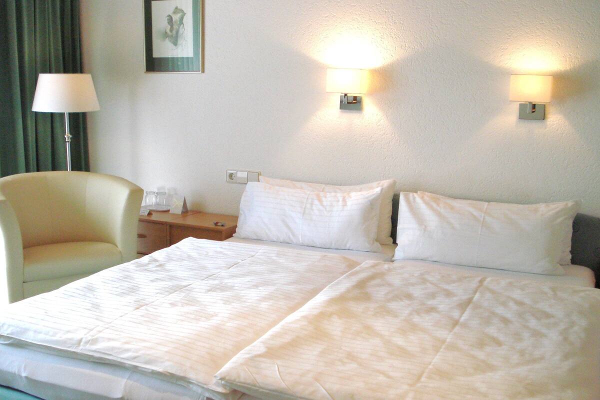 A hotel room with a double bed, a chair, a nightstand, and two wall-mounted lamps.