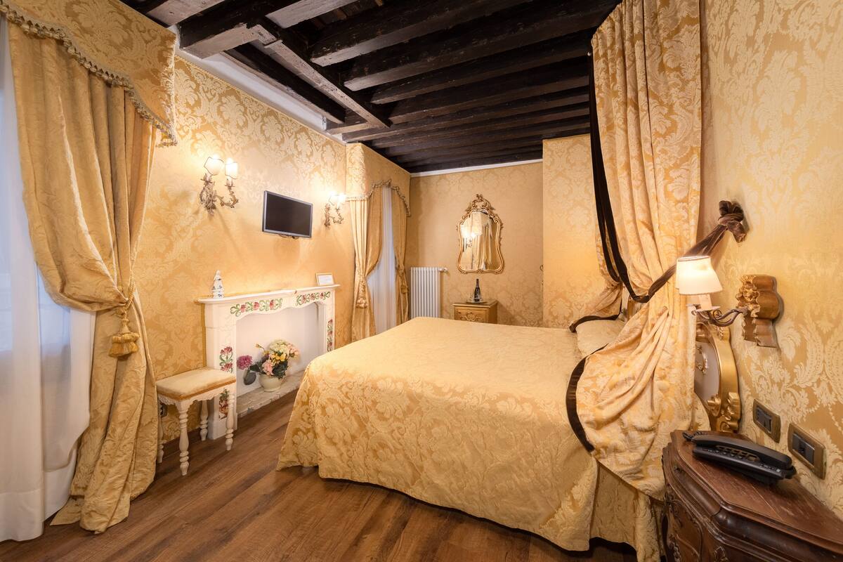 A classic room with a large bed, a wooden floor, a small table with a lamp, and a mirror on the wall.