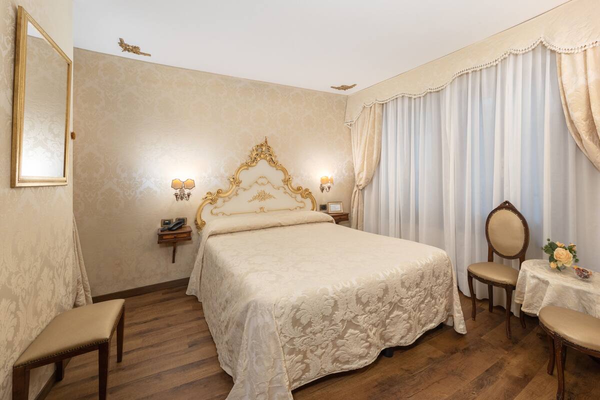 A bedroom with a large bed, a bedside table, a chair, a small table with a vase, and a mirror on the wall.