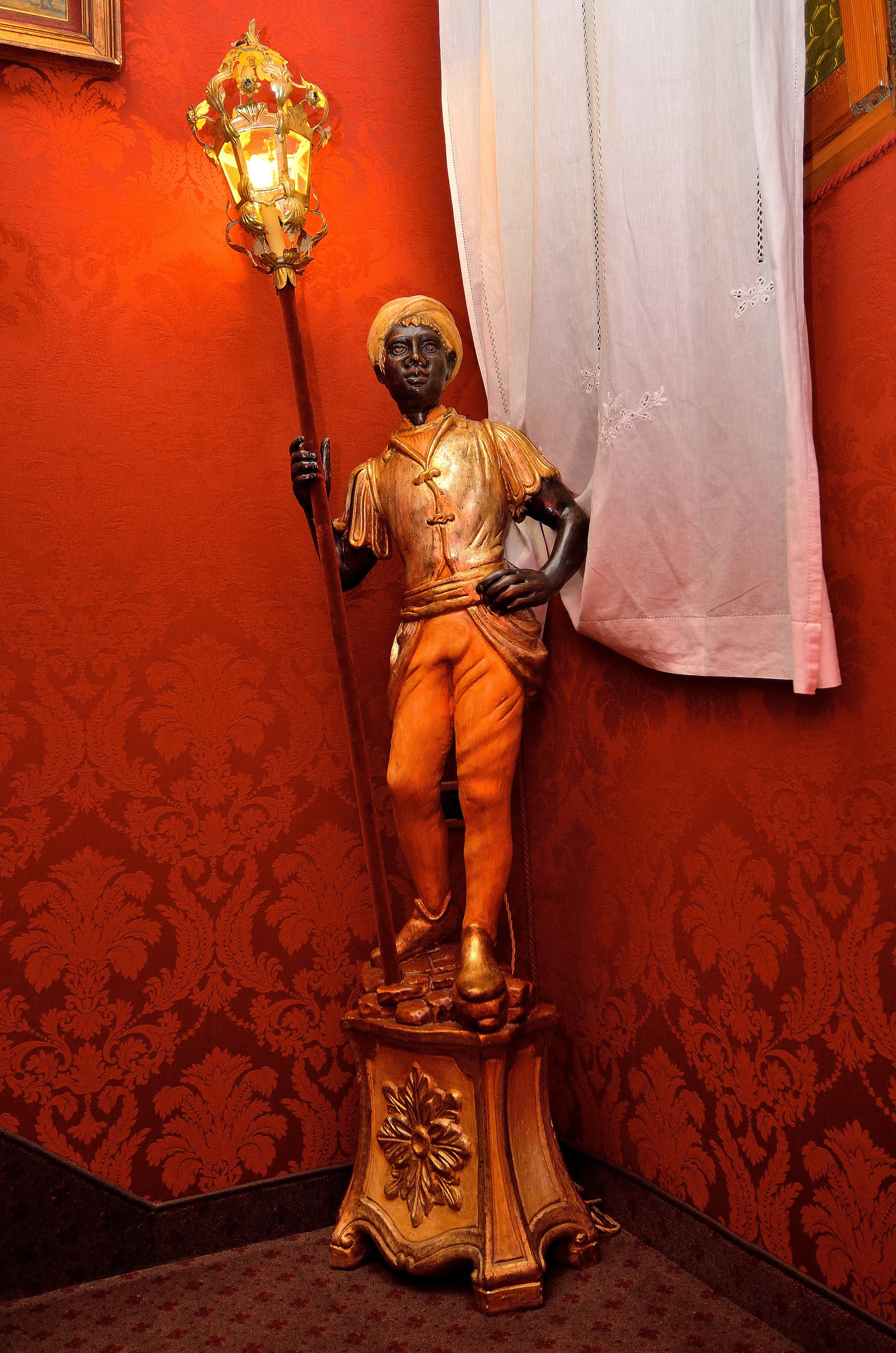 Una statua raffigura una figura che regge uno scettro, posizionata su un piedistallo decorato con motivi floreali, davanti a una parete rossa con un disegno a motivi e una tenda bianca parzialmente visibile.