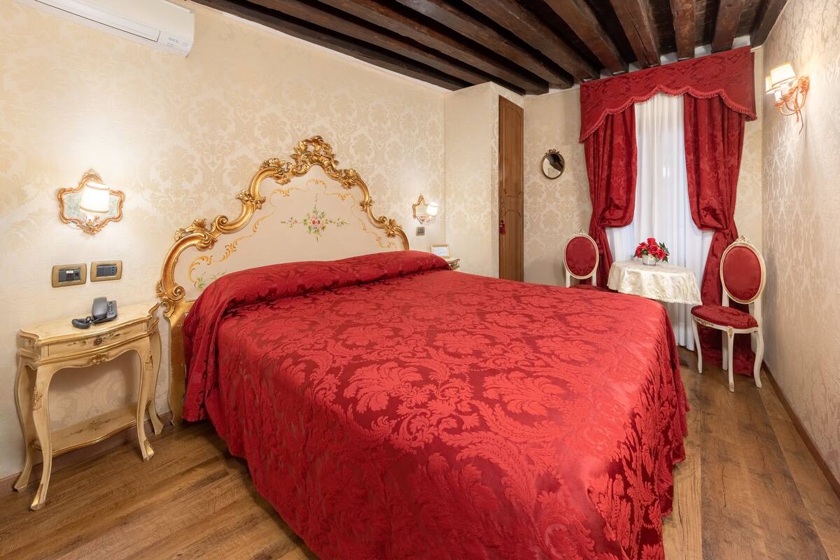 A bedroom with a large bed, a bedside table with a lamp, a chair, and a small table with a vase of flowers.