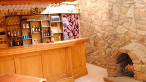 Een bar met houten planken waarop verschillende flessen en glazen staan, een stenen muur en een open haard.