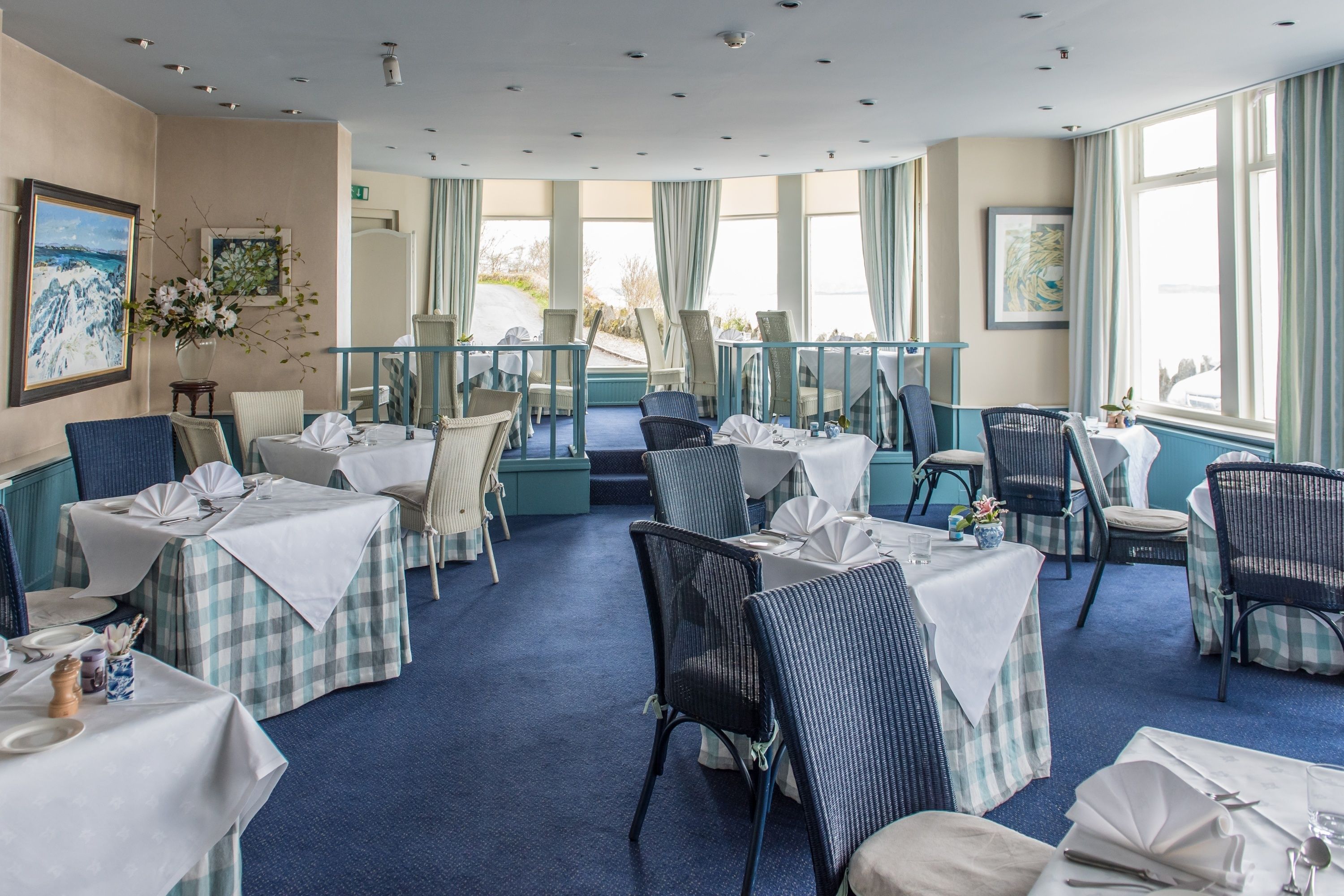 A dining area with checkered tablecloths, wicker chairs, and large windows with curtains.