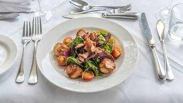 A white plate with a salad of greens, tomatoes, and meat, accompanied by silverware and a glass of water on a white tablecloth.