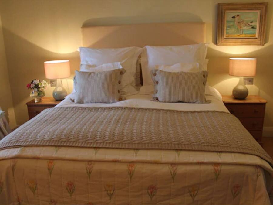 Una cama bien tendida con una colcha de motivos florales y dos almohadas a juego, flanqueada por dos mesitas de noche con lámparas y un jarrón de flores.