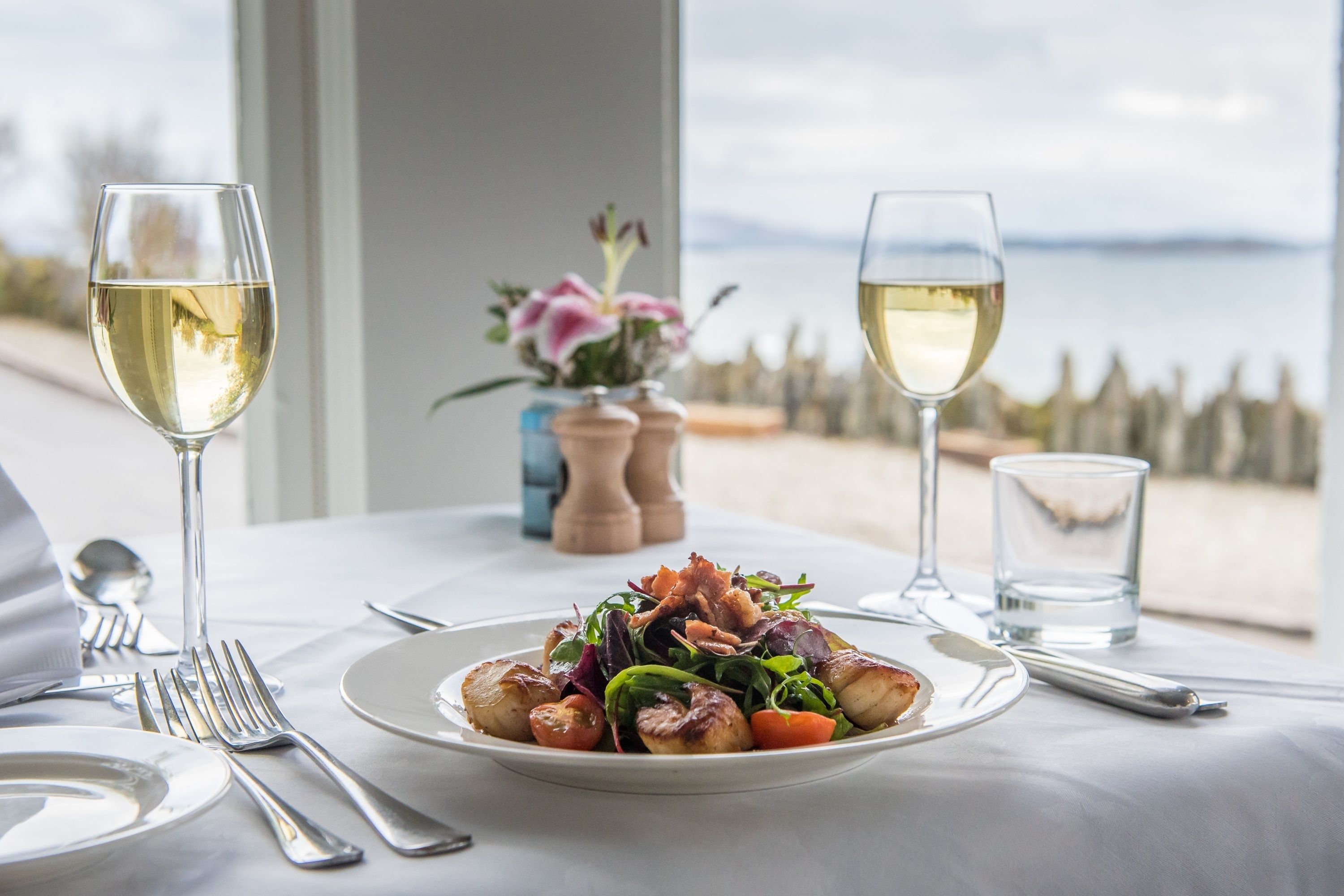 Une table à manger avec une assiette de nourriture, deux verres à vin et une vue sur la mer.