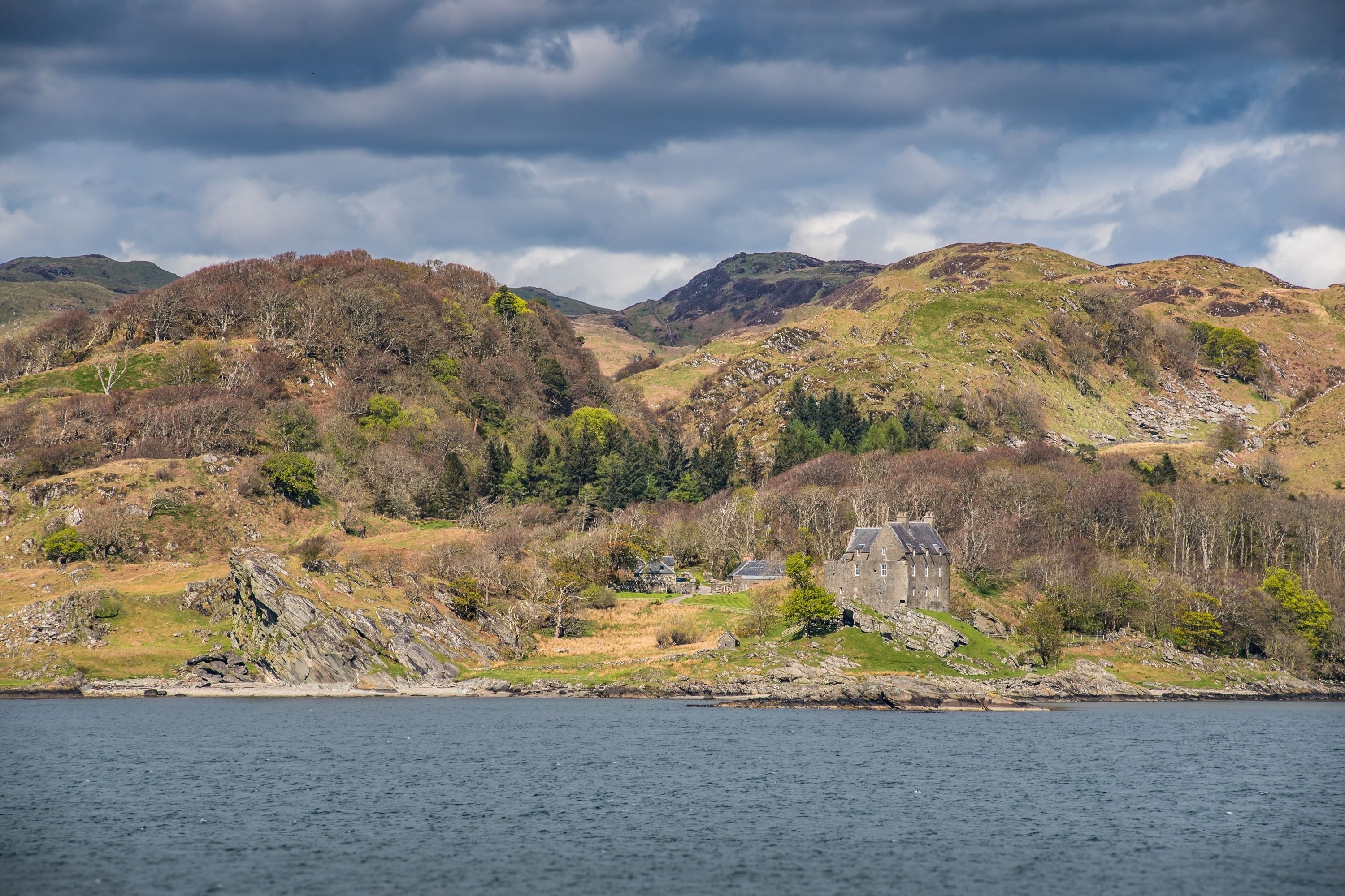 A coastal landscape with a stone building, possibly a hotel, surrounded by hills and a body of water.