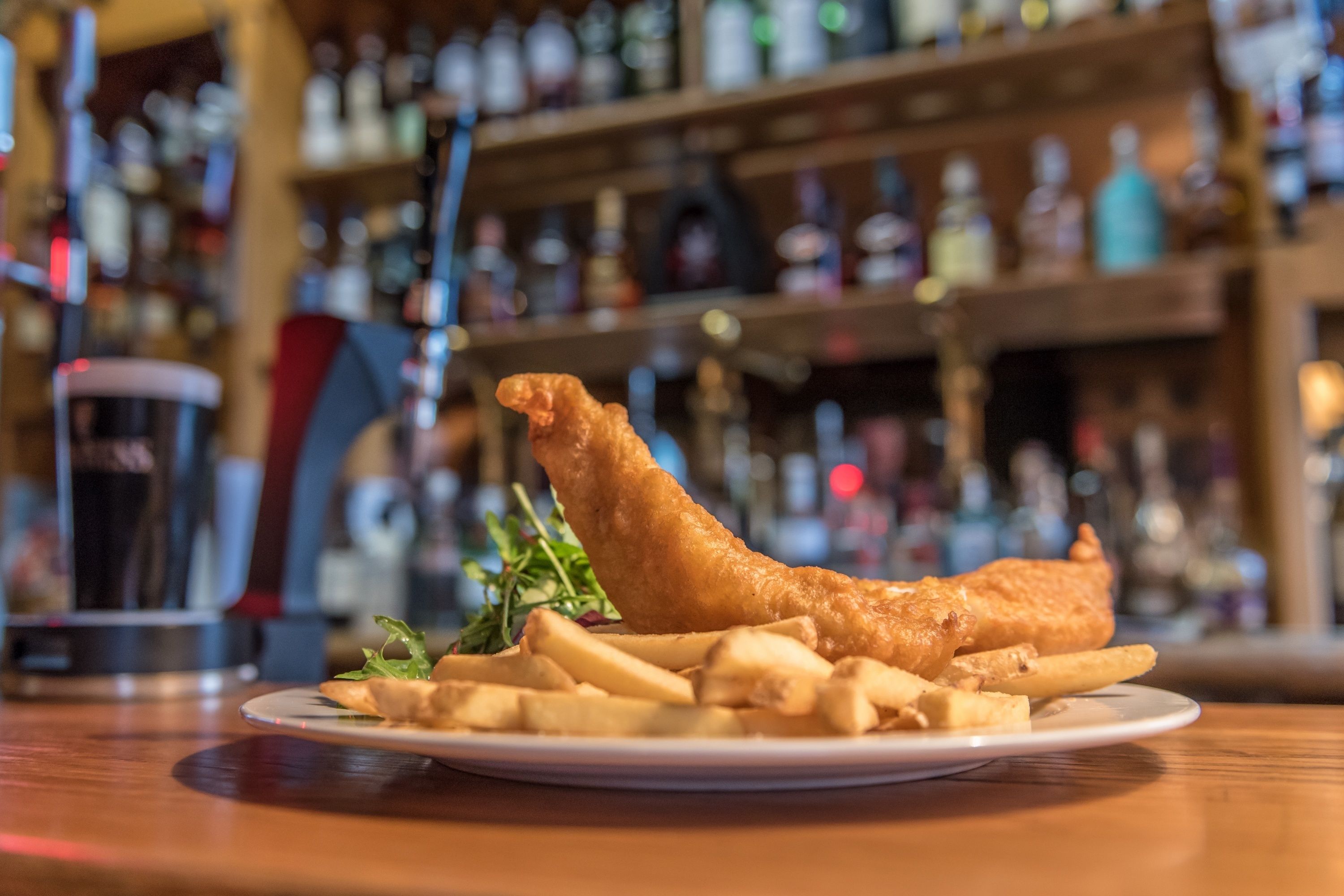 Un plato de pescado con papas fritas y una guarnición de papas y vegetales verdes, sobre una mesa de madera con un fondo borroso de estantes y botellas.