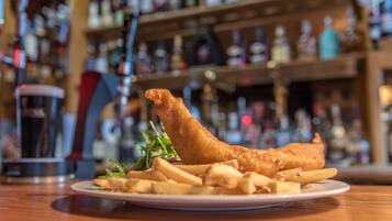Un plato de pescado con papas fritas y una guarnición de papas y vegetales verdes, sobre una mesa de madera con un fondo borroso de estantes y botellas.