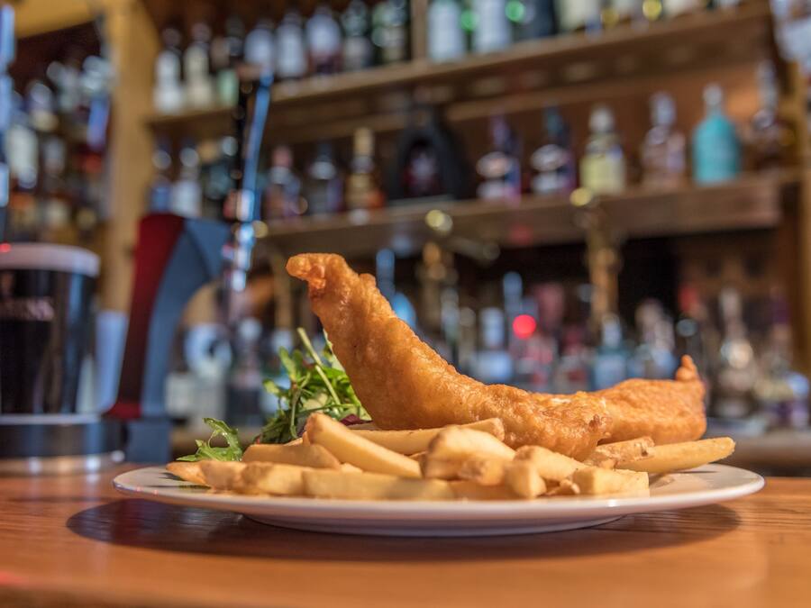 Un plato de pescado con papas fritas y una guarnición de papas y vegetales verdes, sobre una mesa de madera con un fondo borroso de estantes y botellas.