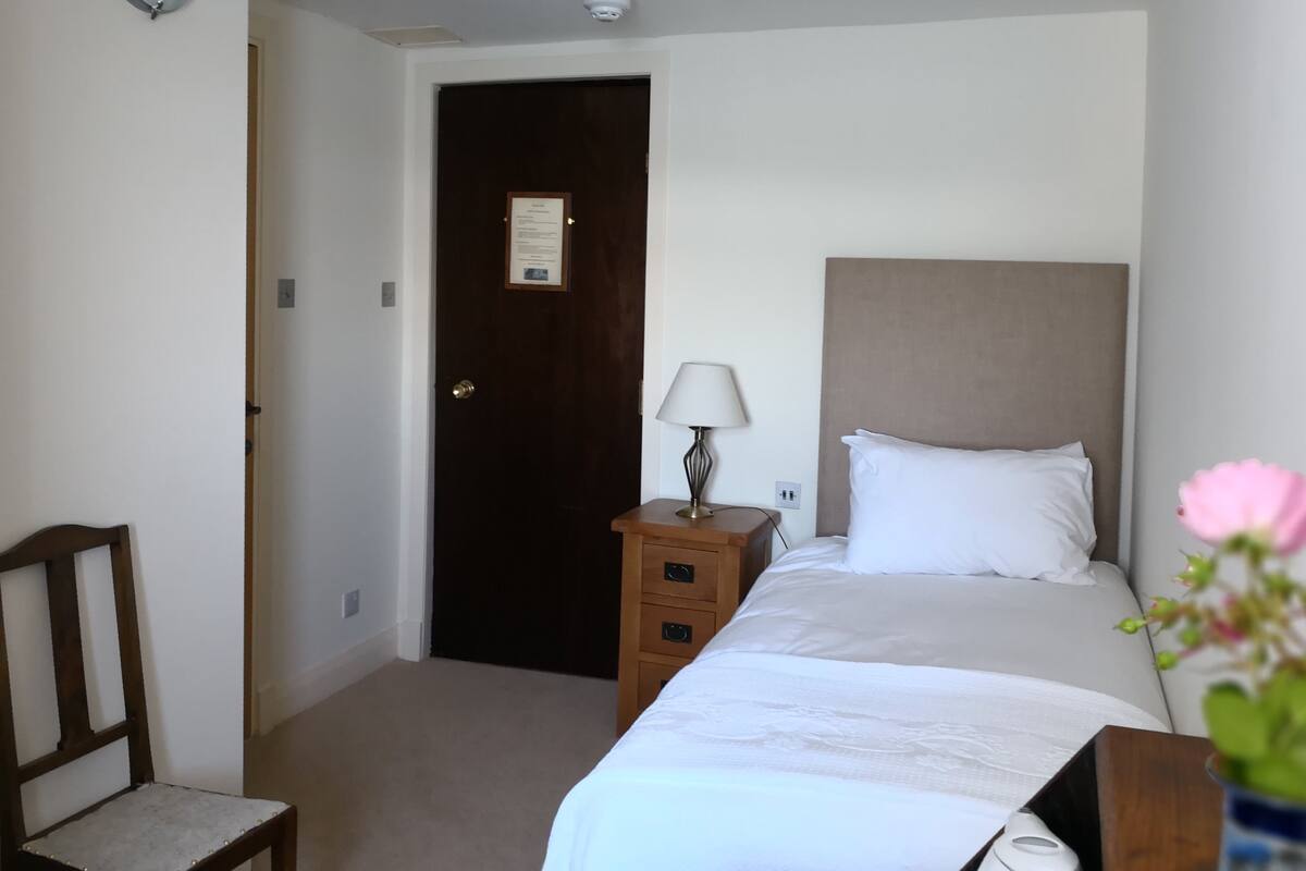 Quarto de hotel com uma cama, uma cadeira, mesa de cabeceira com abajur e uma porta com aviso.