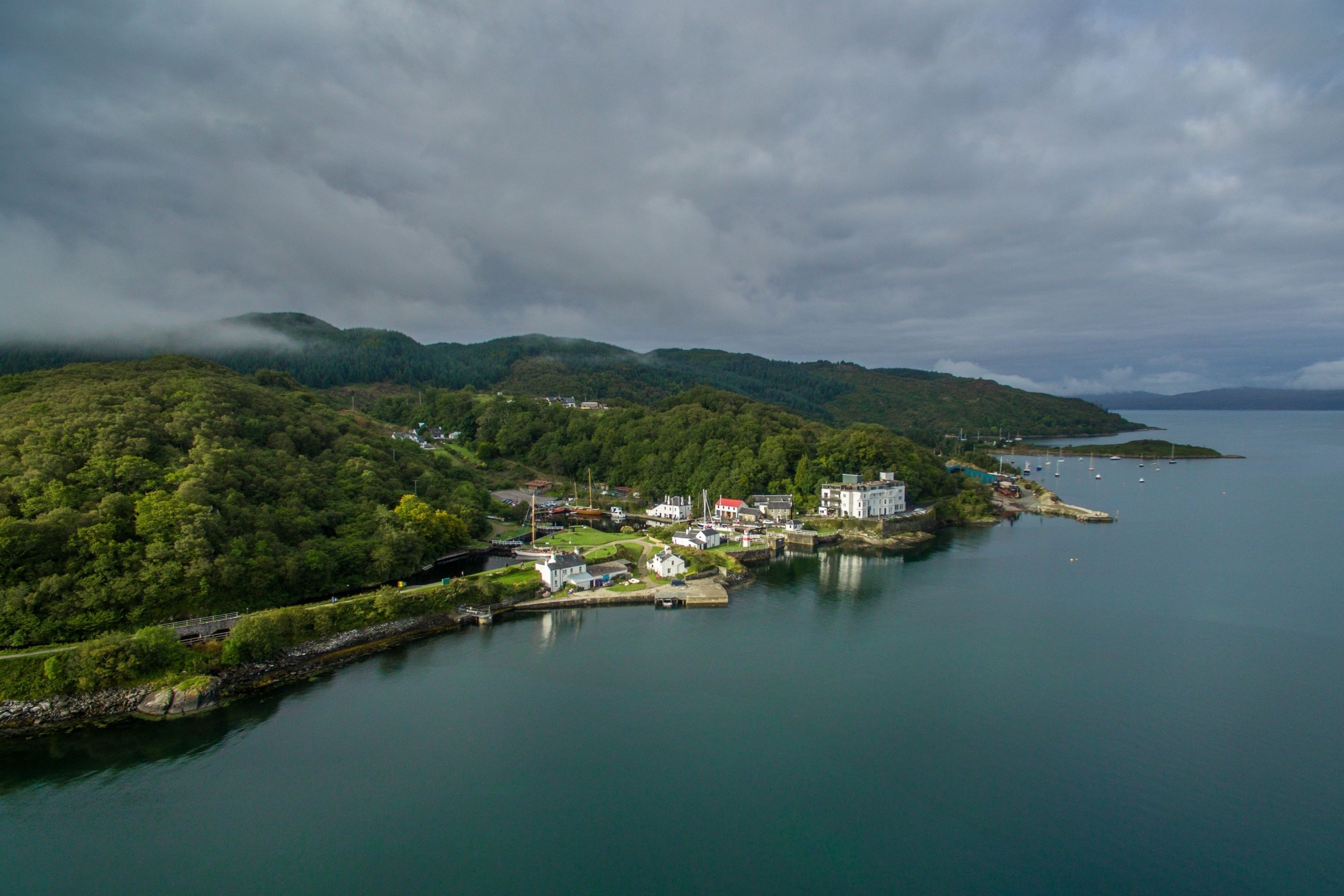 A coastal town with a marina, surrounded by lush green hills and a calm sea.