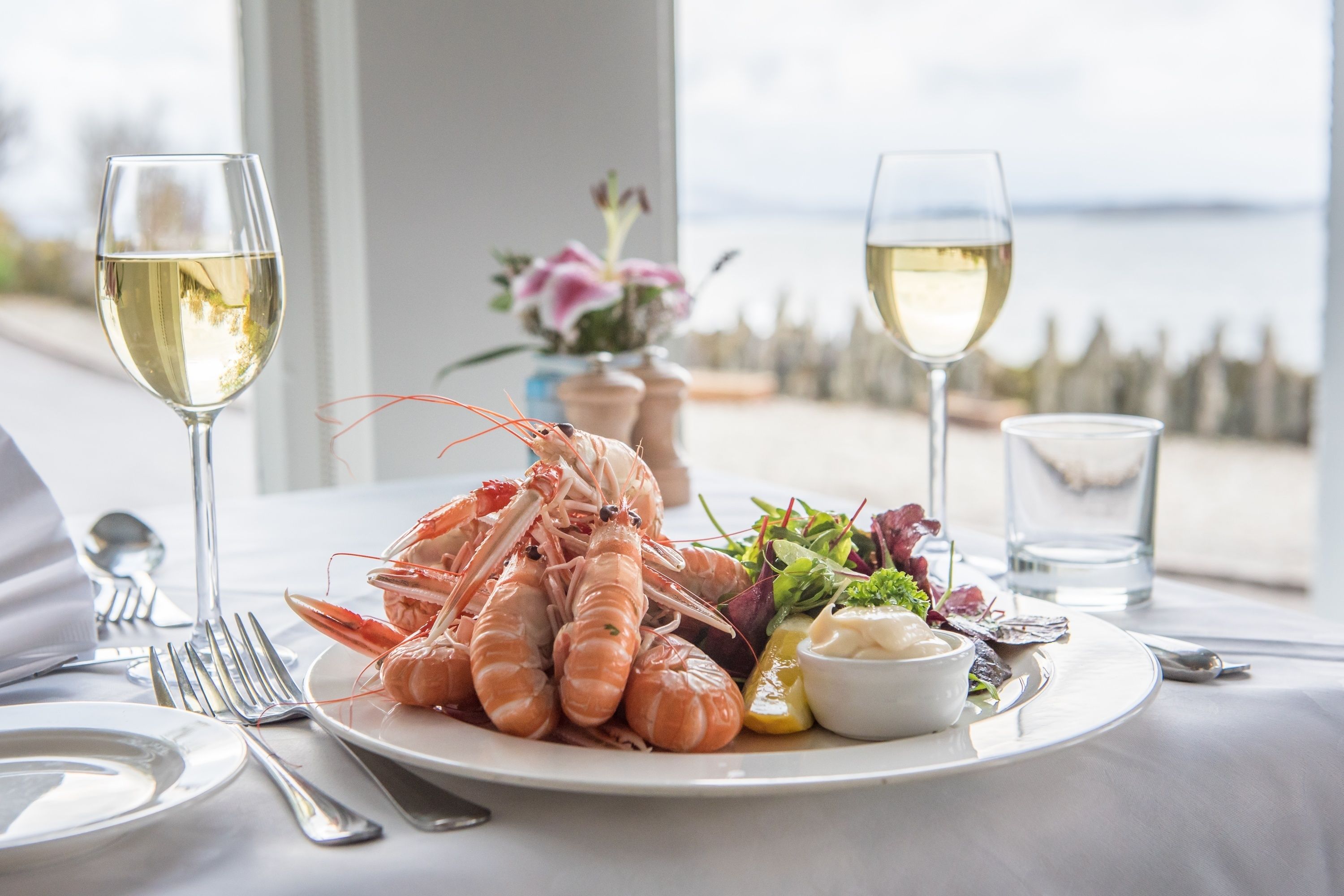 Plato de mariscos con camarones, ensalada y limón, acompañado de dos copas de vino blanco, sobre una mesa con vista al mar.