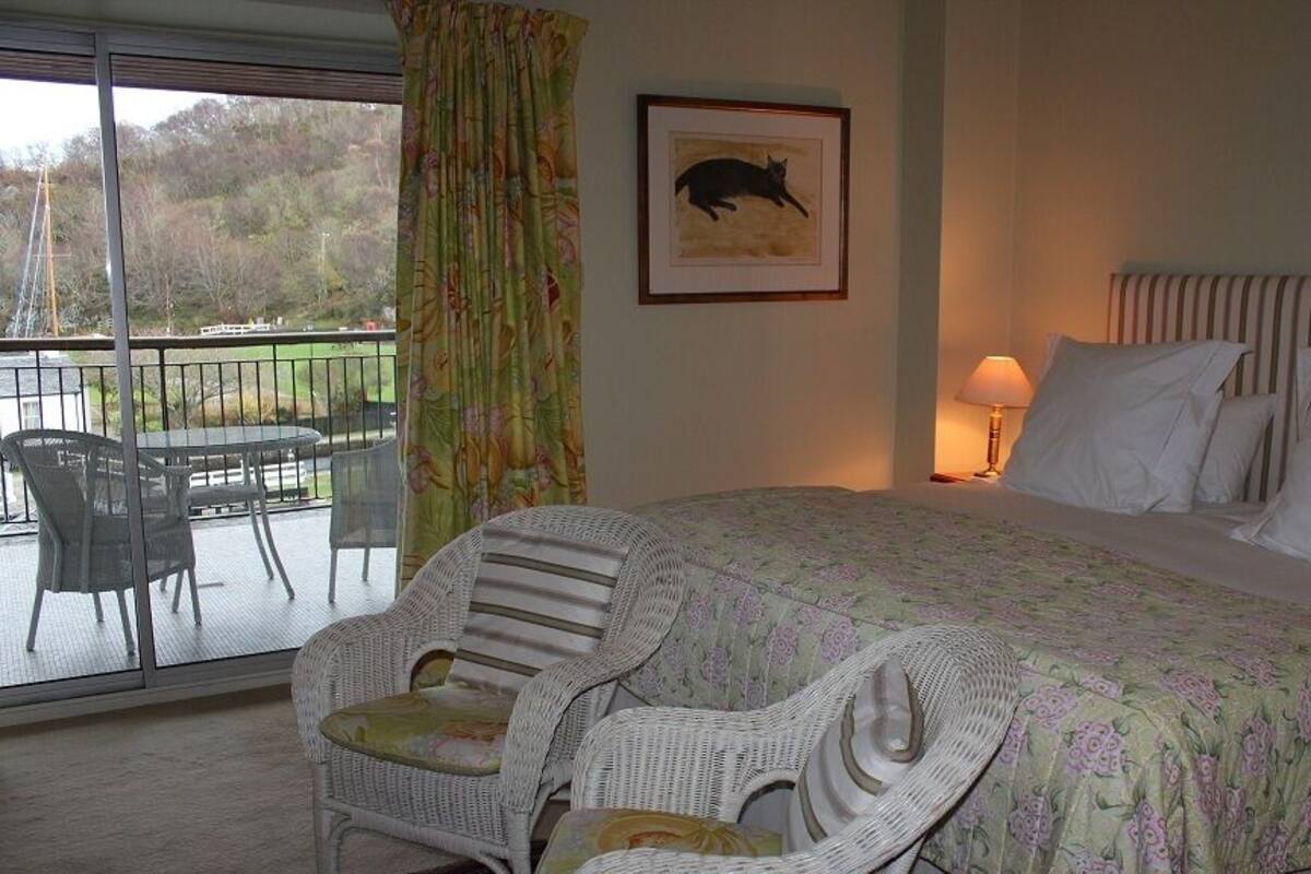Quarto com cama, duas cadeiras, janela com vista para um corpo d'água e árvores, e um quadro na parede.