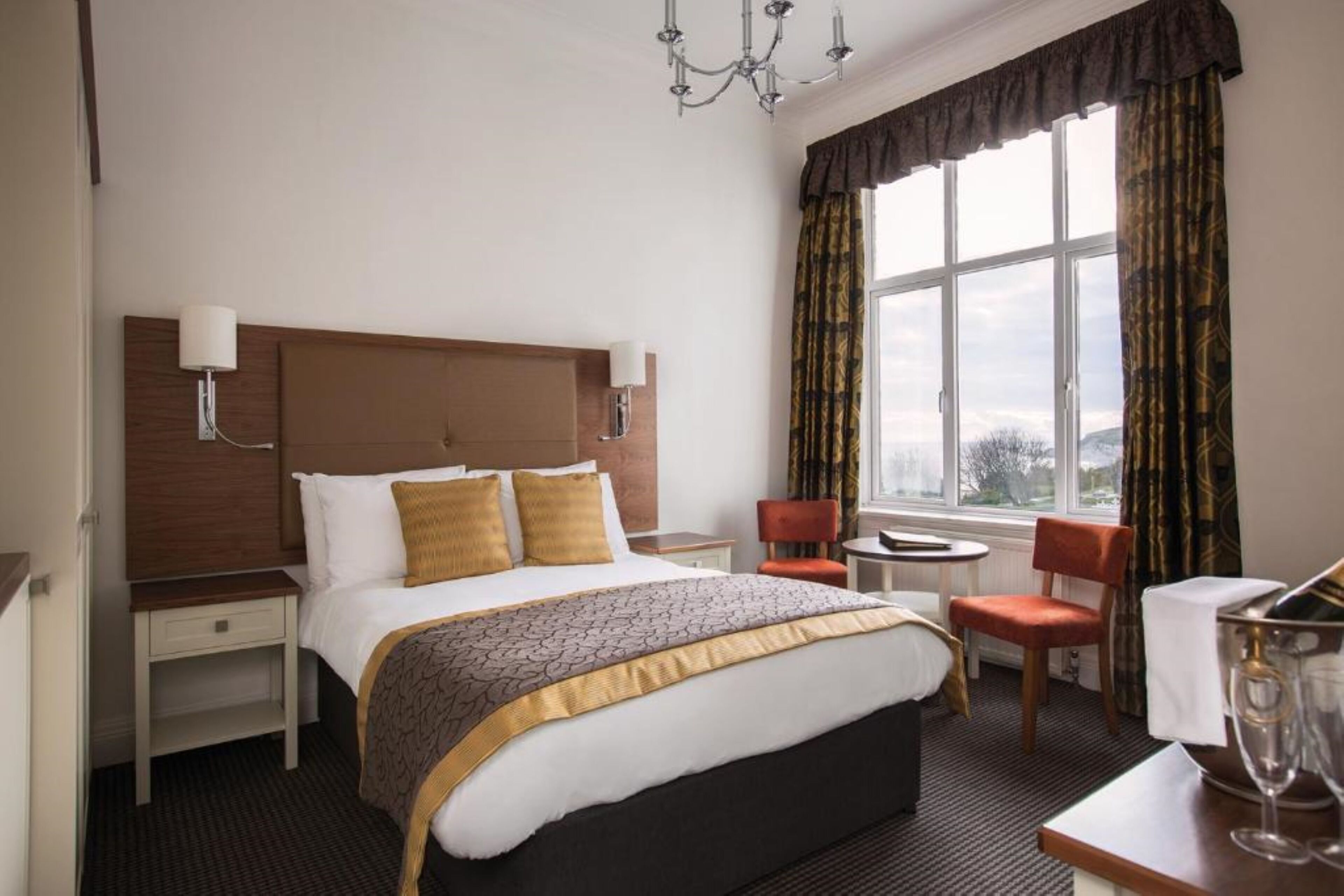 A hotel room with a large bed, a wooden headboard, two bedside tables, a small round table, two chairs, a chandelier, and a view through the window.