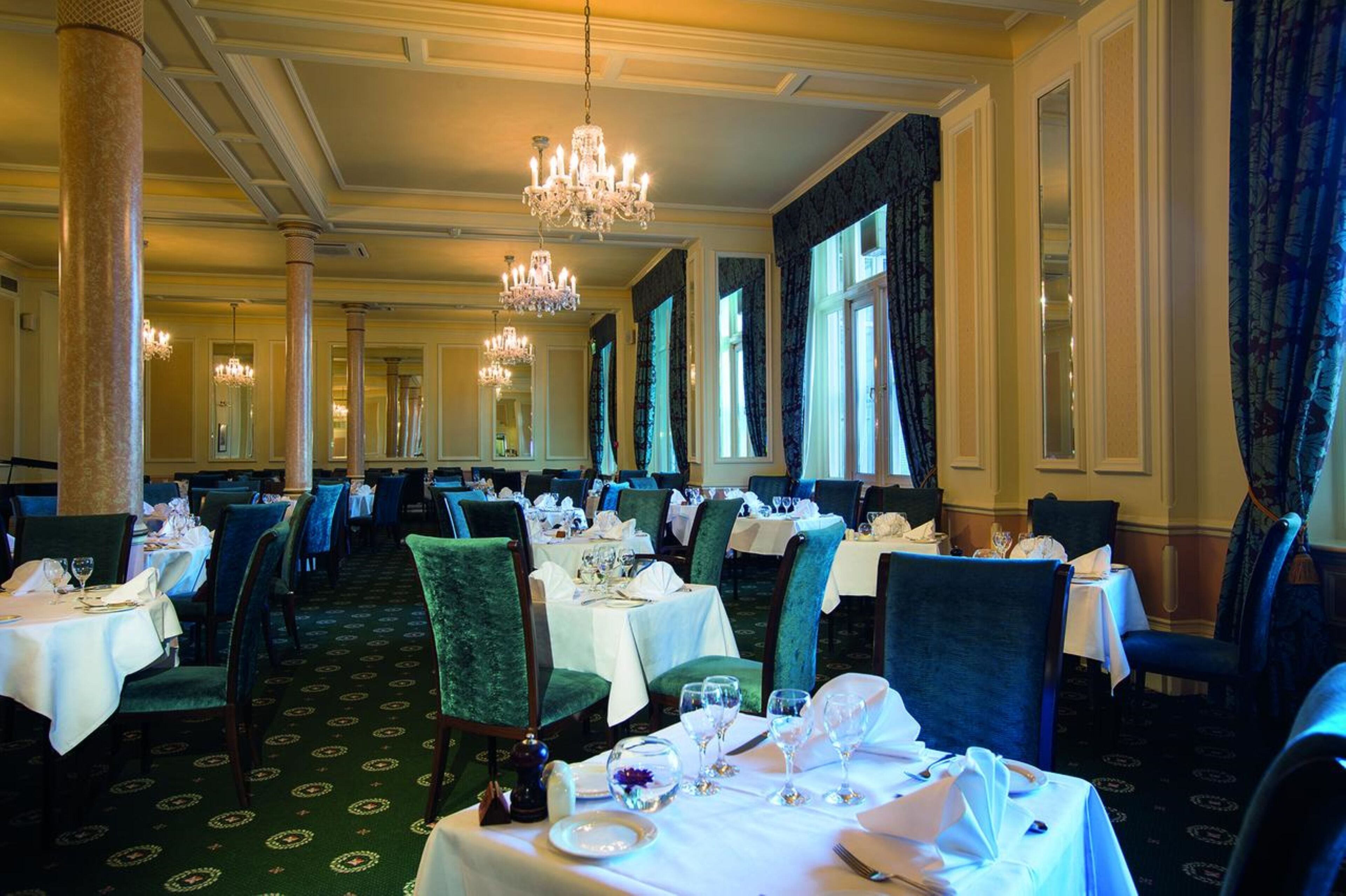 Un comedor formal con varias mesas dispuestas para el servicio, amplias ventanas con cortinas y candelabros.