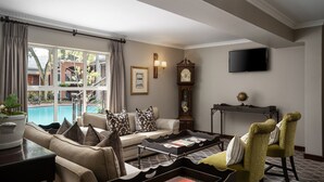 A well-lit living room with a sofa, chairs, a coffee table, and a television. A large window offers a view of a swimming pool and outdoor area. - Premier Hotel Falstaff (Sandton)