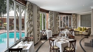 A dining area with tables set for four, large windows with curtains, a pool view, and a sofa with cushions. - Premier Hotel Falstaff (Sandton)