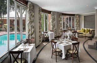 Comedor con mesas dispuestas para cuatro, ventanas amplias con cortinas, vista a la piscina y un sofá con cojines.