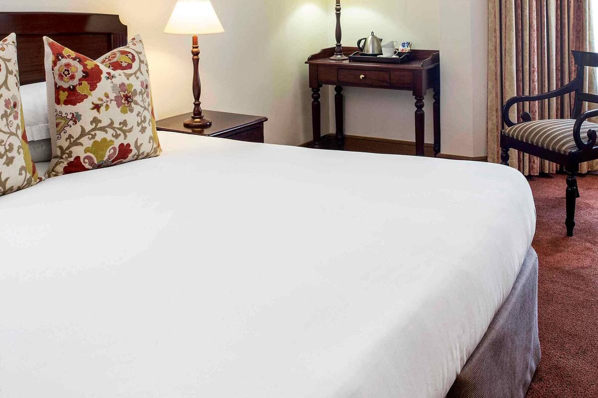 Een hotelkamer met een groot bed, twee bedlampen, een klein tafeltje met lamp, een stoel en lijstjes met kunstwerken aan de muren.