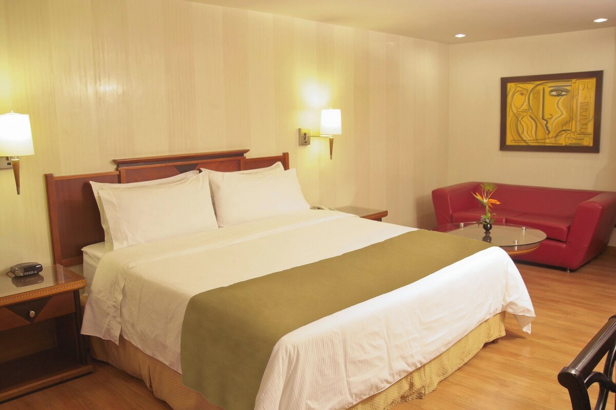 Een hotelkamer met een bed, nachtlampjes, een rode bank, een kleine tafel en een aan de muur hangend schilderij.