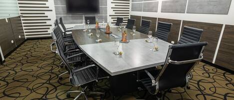 A conference room with a long table, black chairs, and a large screen on the wall.