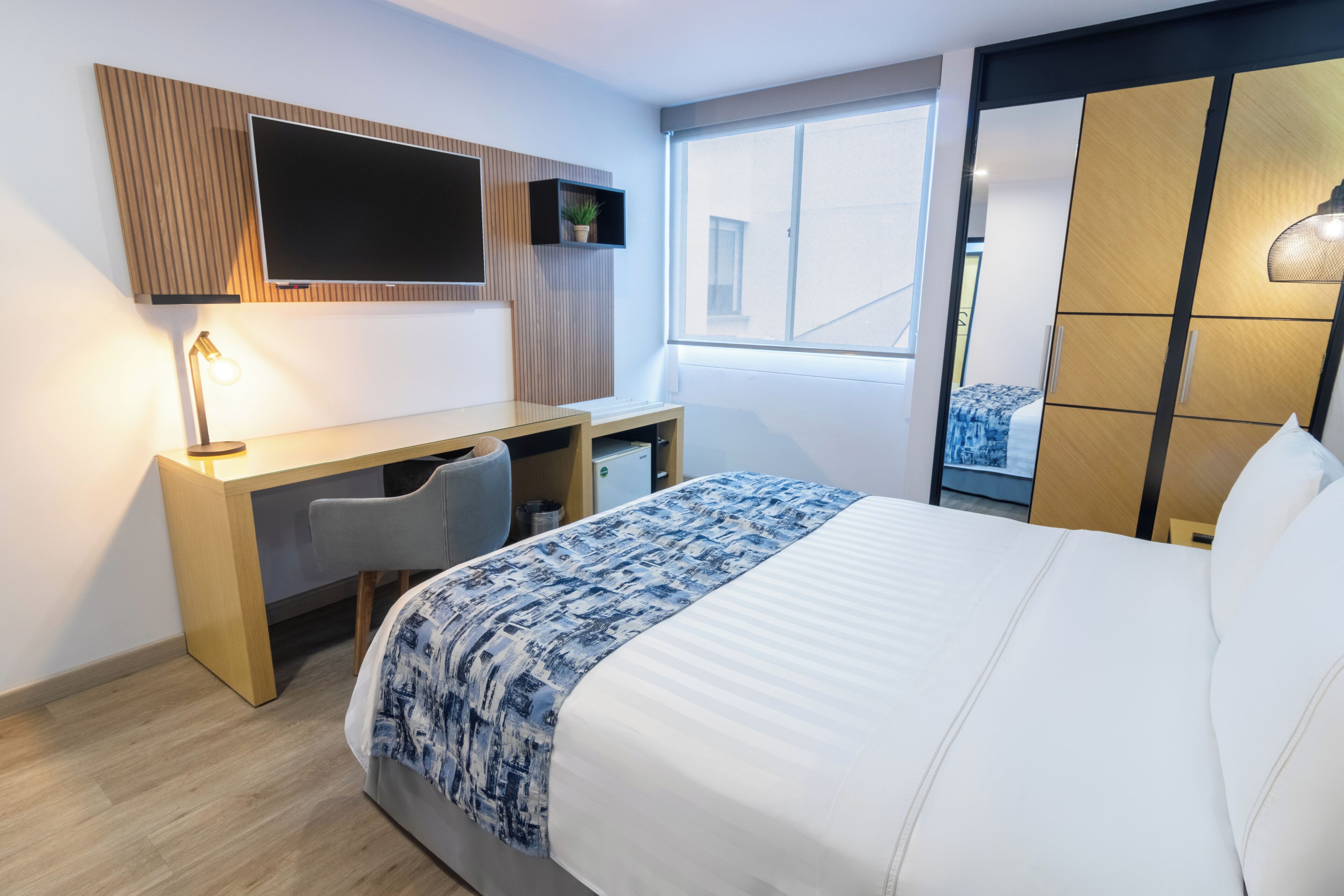 Una camera d'albergo con un letto, una scrivania, una sedia, una TV a parete, una finestra e un armadio.