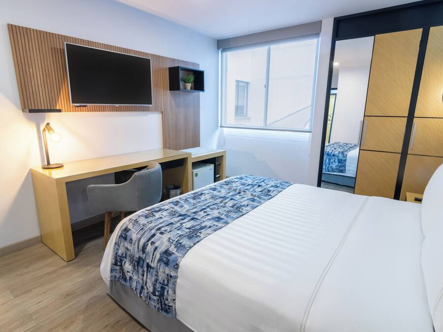 Habitación de hotel con cama, escritorio, silla, televisor montado en la pared, ventana y armario.