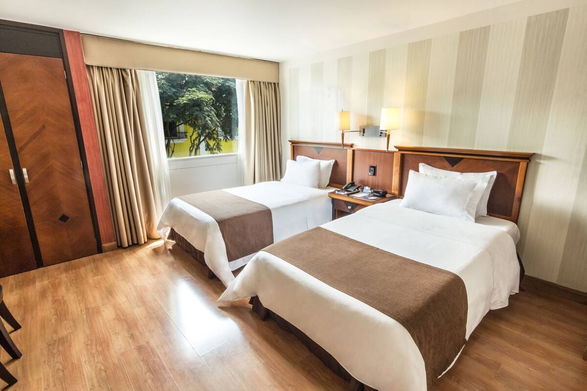 A hotel room with two beds, a wooden floor, a window with curtains, a nightstand with a lamp, and a wall-mounted phone.