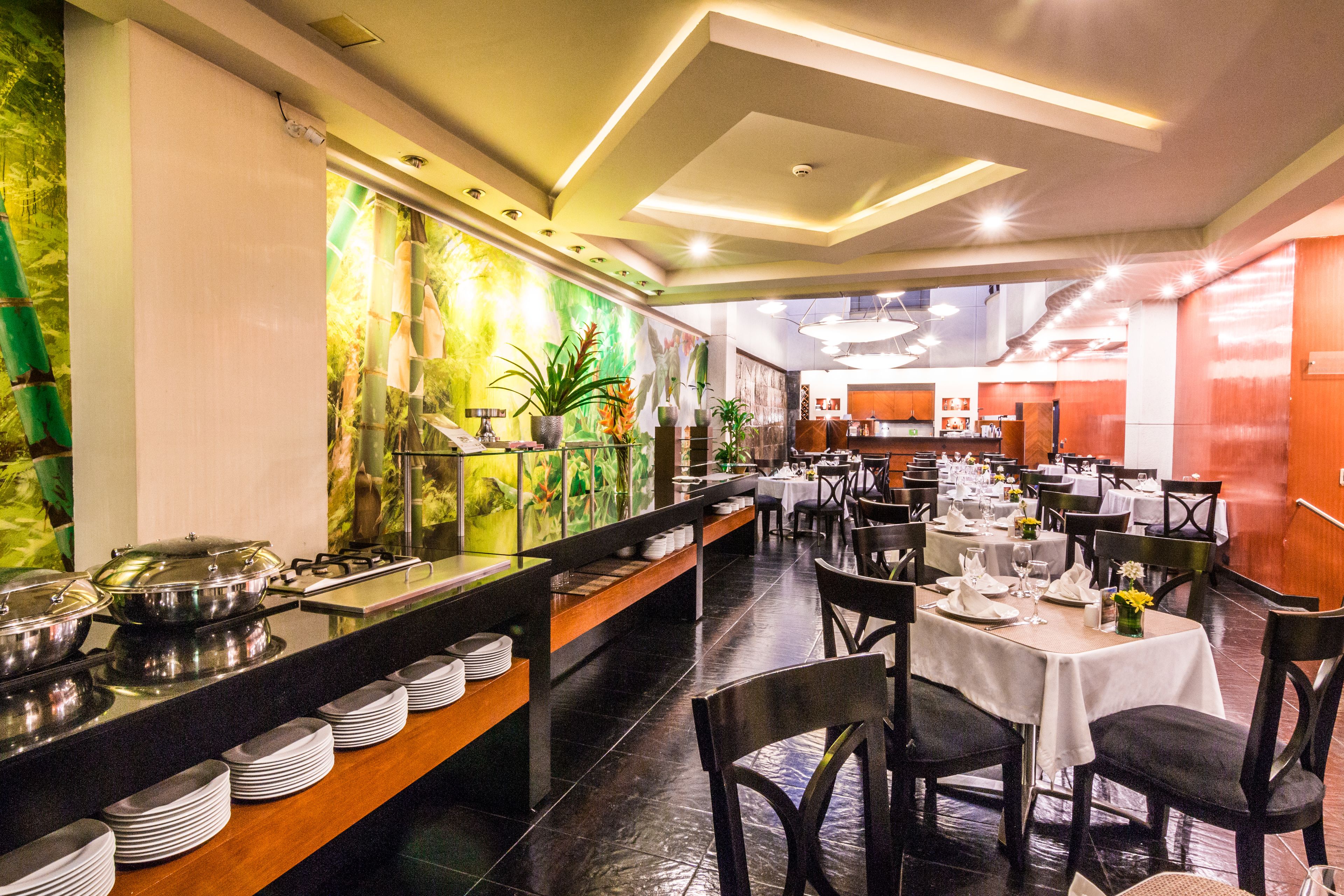 Un restaurant proposant un buffet avec plusieurs points de service, des tables dressées et une variété de plantes et d’œuvres d’art disposées sur les murs.