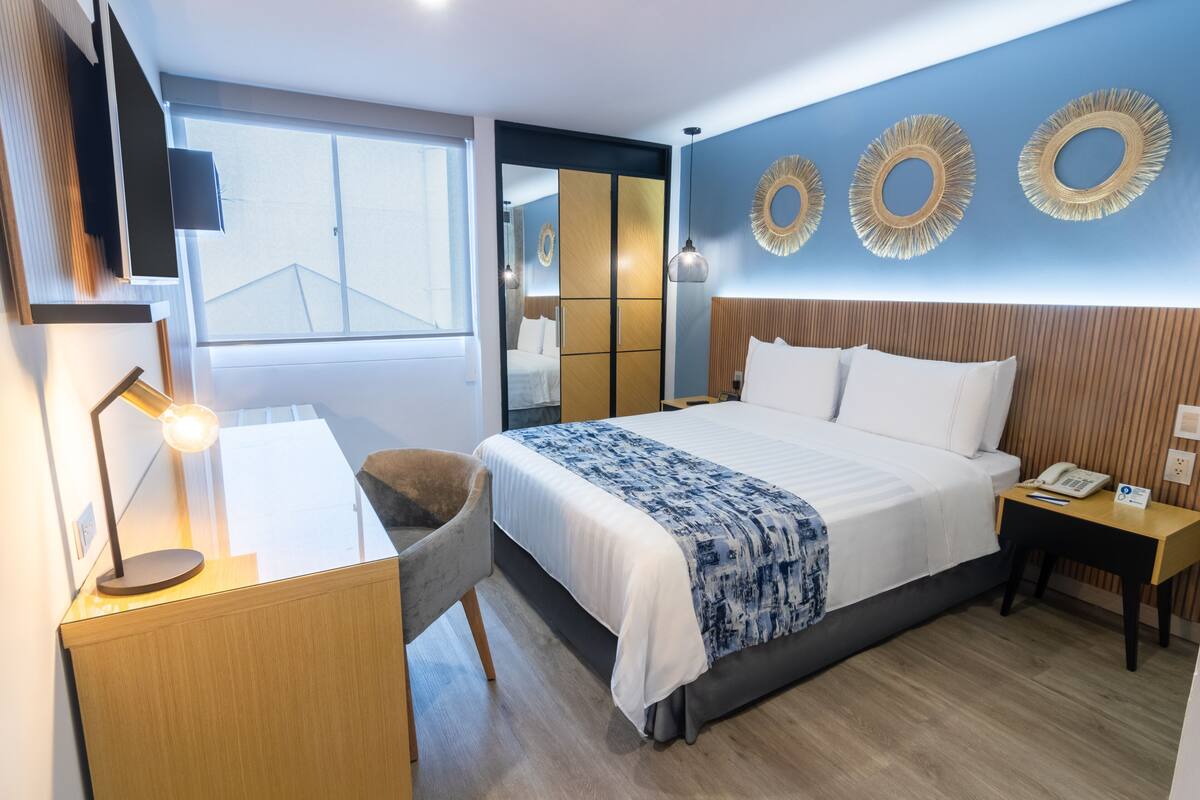 A modern hotel room with a large bed, a desk with a lamp, a chair, and a bedside table with a phone.