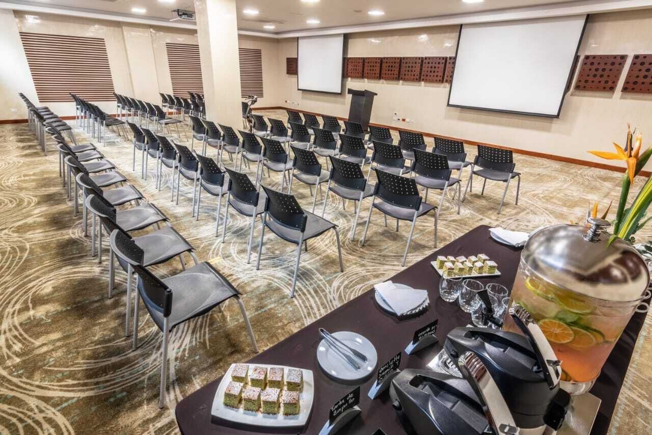 A conference room with rows of chairs, a projector screen, a podium, and a buffet table with food and a drink dispenser.