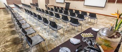 A conference room with rows of chairs, a projector screen, a podium, and a buffet table with food and a drink dispenser.