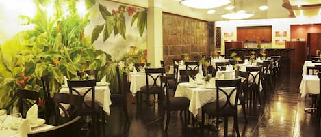 Restaurante com várias mesas e cadeiras, um grande mural com plantas, e uma área de cozinha ao fundo.
