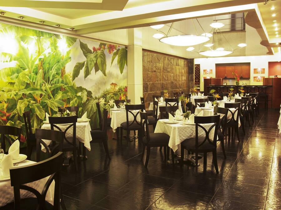 A restaurant with multiple tables and chairs, a large mural with plants, and a kitchen area in the background.