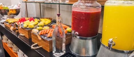Un bufé con diversas opciones de comida y bebidas, que incluye una sección de frutas, una barra de jugos y una presentación de cereales.