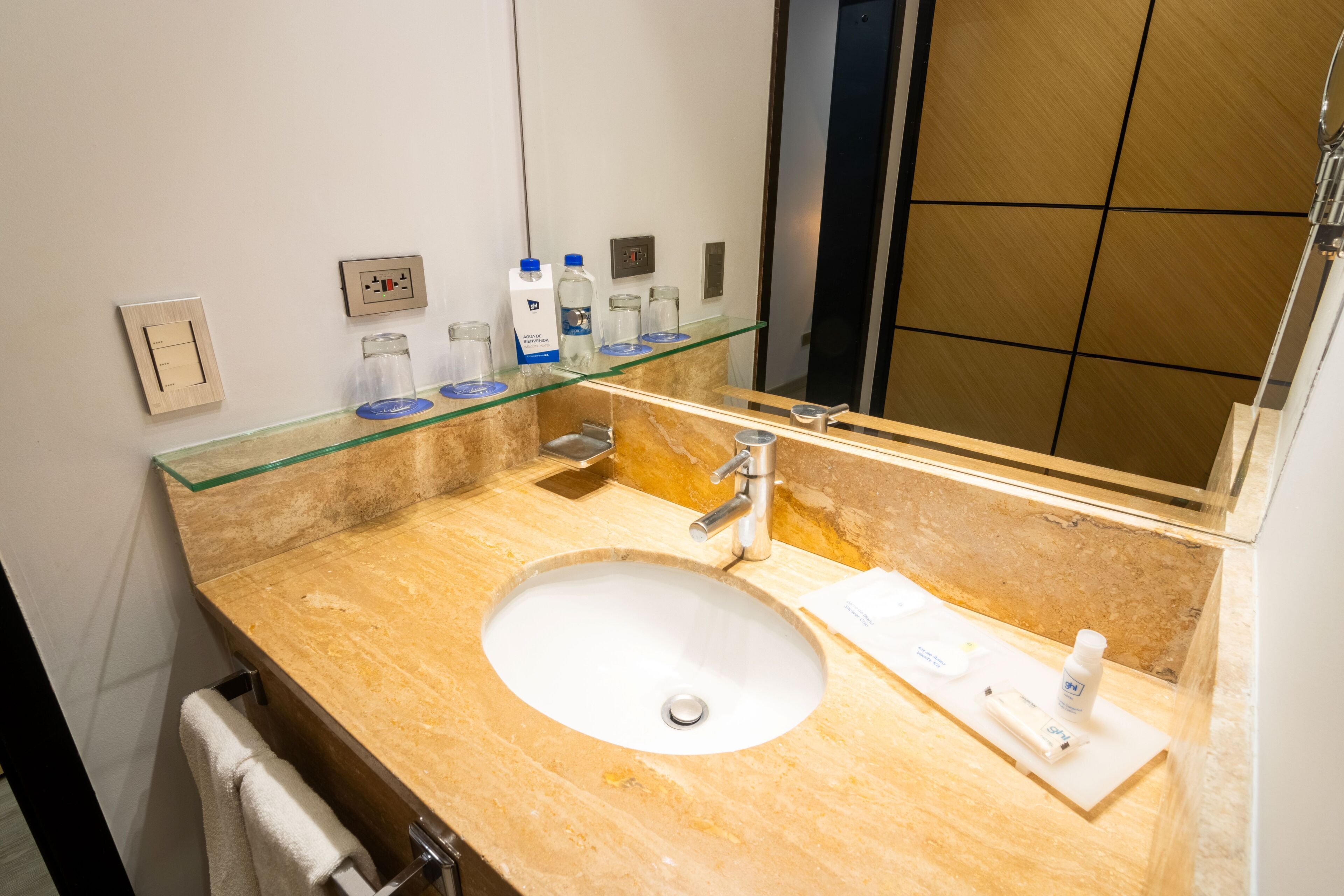 A hotel bathroom with a marble sink, a glass shelf with toiletries, a mirror, and a towel rack.