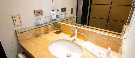 A hotel bathroom with a marble sink, a glass shelf with toiletries, a mirror, and a towel rack.