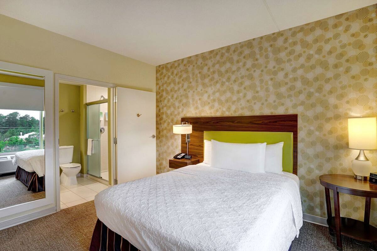 A hotel room with a bed, a bedside table with a lamp, a bathroom with a toilet and shower, and a view of the outdoors.