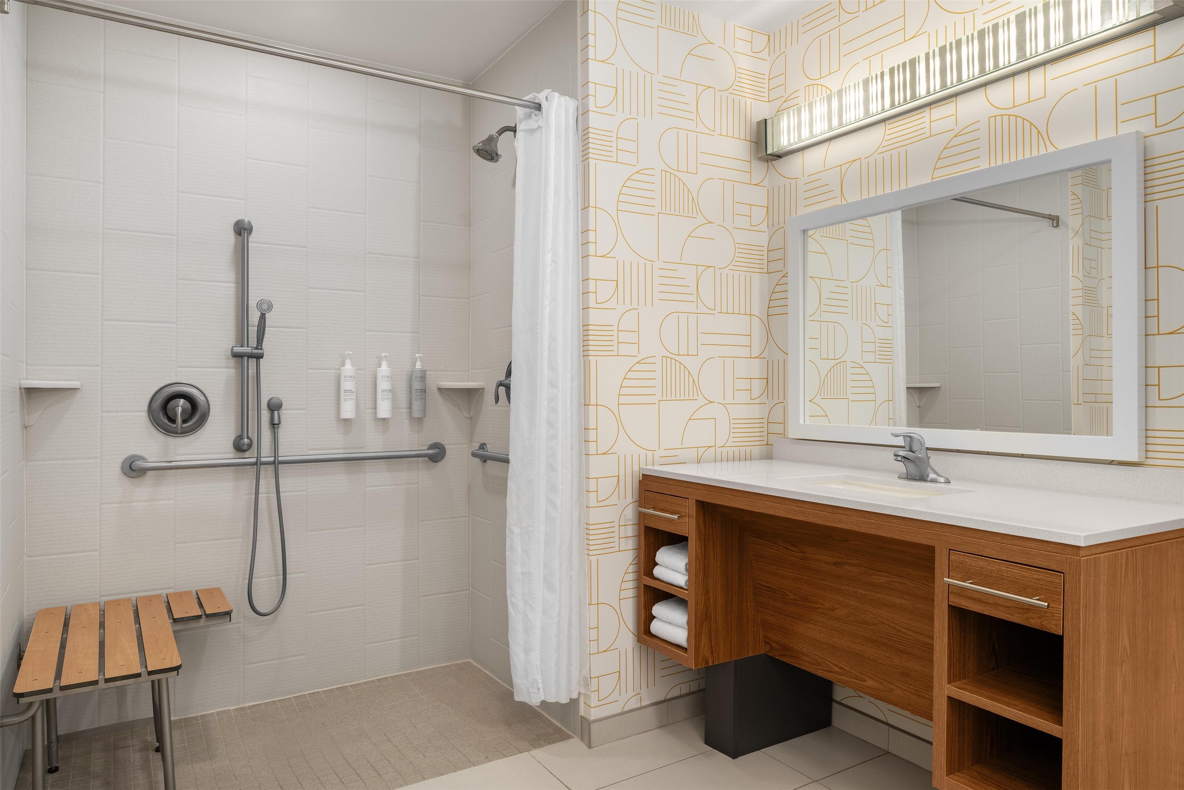 A modern bathroom with a shower, a wooden bench, a vanity with a mirror, and patterned wall tiles.