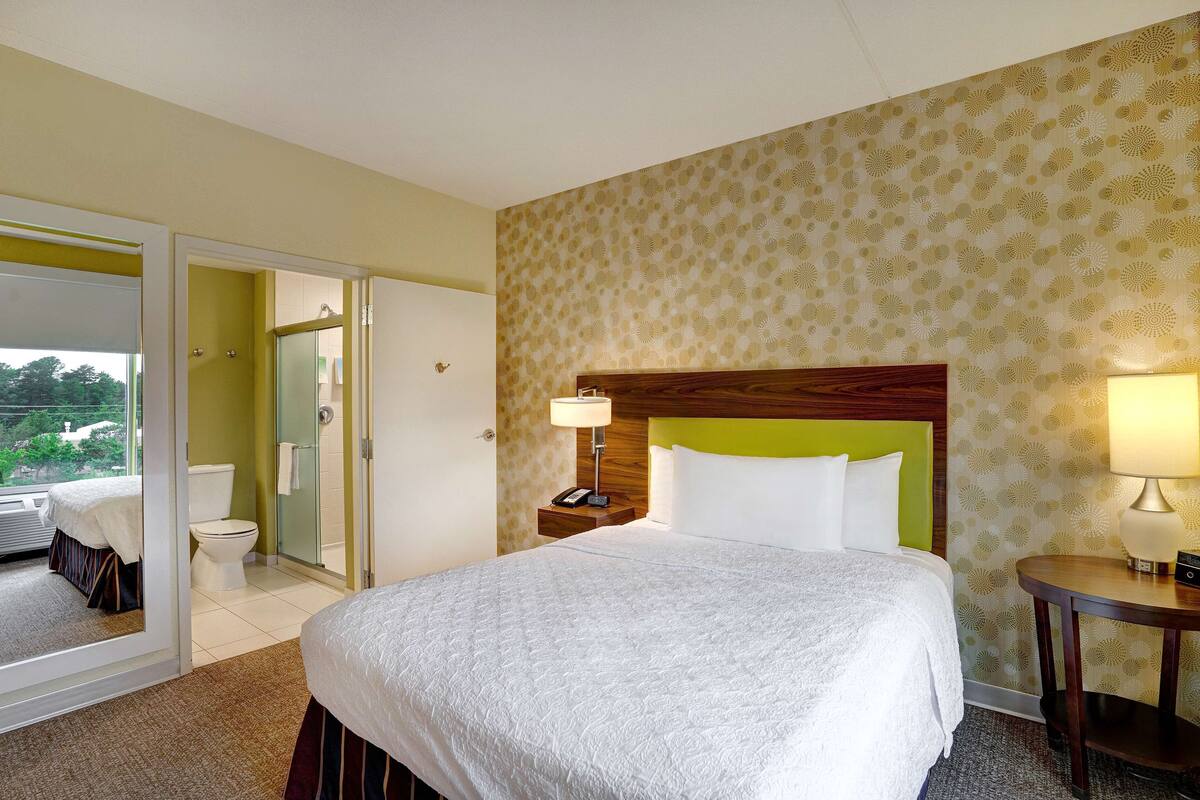 A hotel room with a bed, a bedside table with a lamp, a bathroom with a toilet and shower, and a view through an open door.