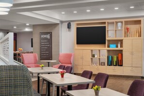 A hotel lobby with seating, a TV screen, and a sign about a home guarantee. - Home2 Suites by Hilton Lexington Park Patuxent River NAS, MD (Lexington Park)