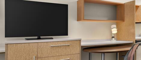 A modern hotel room with a flat-screen TV, wooden cabinets, a desk with a chair, and a bedside lamp.