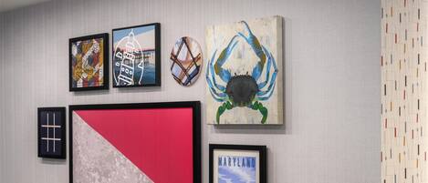 A wall with various framed artworks, including a large red and white piece, a framed crab painting, and a framed sign that says "MARYLAND Welcome to Adventure".