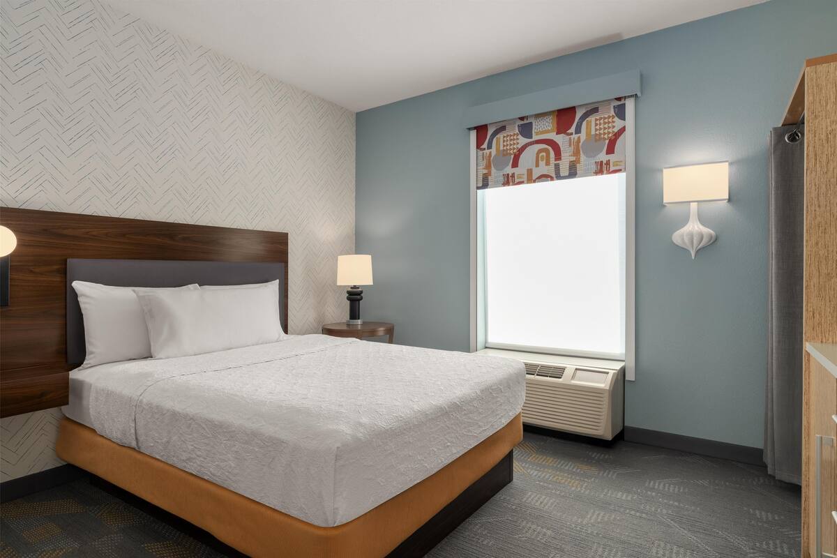 A neatly arranged hotel room with a large bed, a window with a view, a wall-mounted air conditioner, and a nightstand with a lamp.