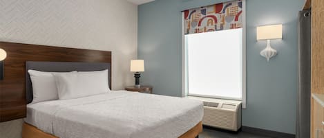 A neatly arranged hotel room with a large bed, a window with a view, a wall-mounted air conditioner, and a nightstand with a lamp.