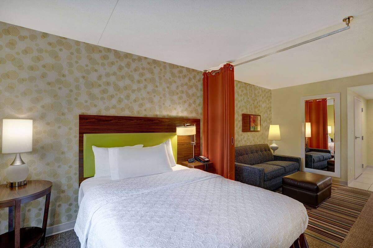 A hotel room with a bed, two chairs, a nightstand, a mirror, and a door.