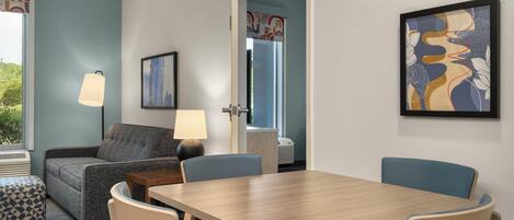 Habitación de hotel con sofá, una mesa, sillas, lámpara, un cuadro enmarcado y ventana con vistas.