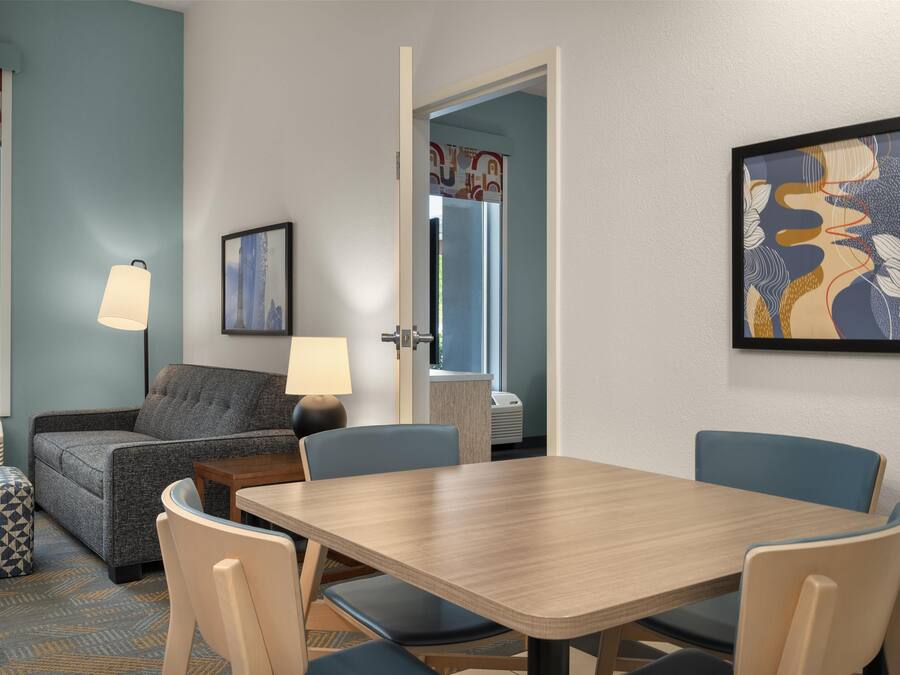 Una camera d'hotel con un divano, un tavolo, sedie, una lampada, un quadro incorniciato e una finestra vista.