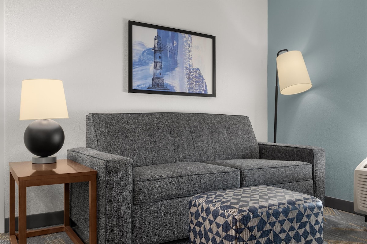 A grey sofa, a wooden side table with a lamp, a patterned ottoman, and a framed picture of a lighthouse on the wall.